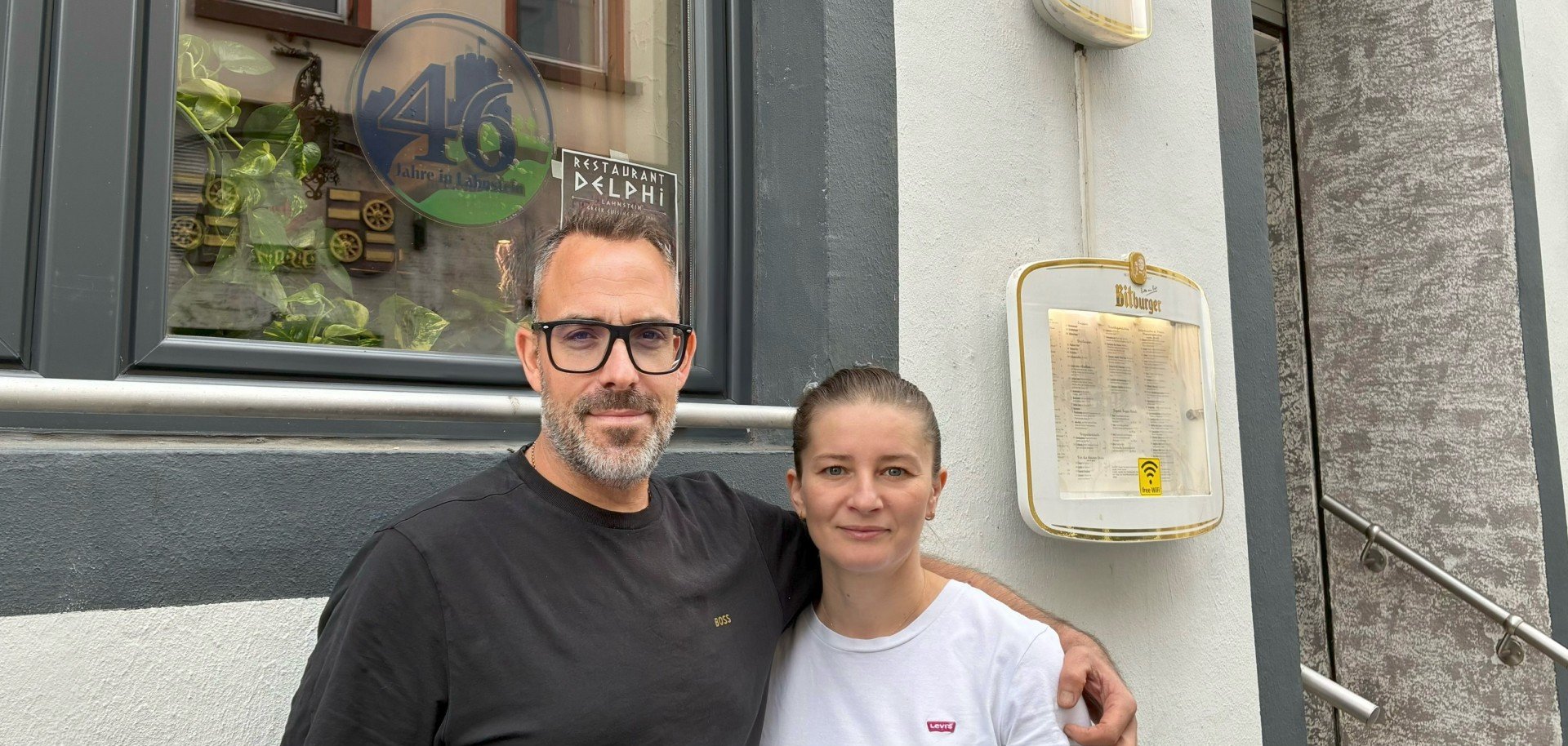 Ein Mann und eine Frau stehen nebeneinander vor der Fassade eines Restaurants und blicken in die Kamera. Der Mann trägt eine schwarze Brille und hat einen Arm um die Frau gelegt. Hinter ihnen ist ein Fenster zu sehen, in dem unter anderem ein Aufkleber mit der Aufschrift „Restaurant Delphi“ angebracht ist. Rechts neben den beiden hängen ein Bitburger-Schild und ein Schaukasten mit der Speisekarte an der Hauswand.