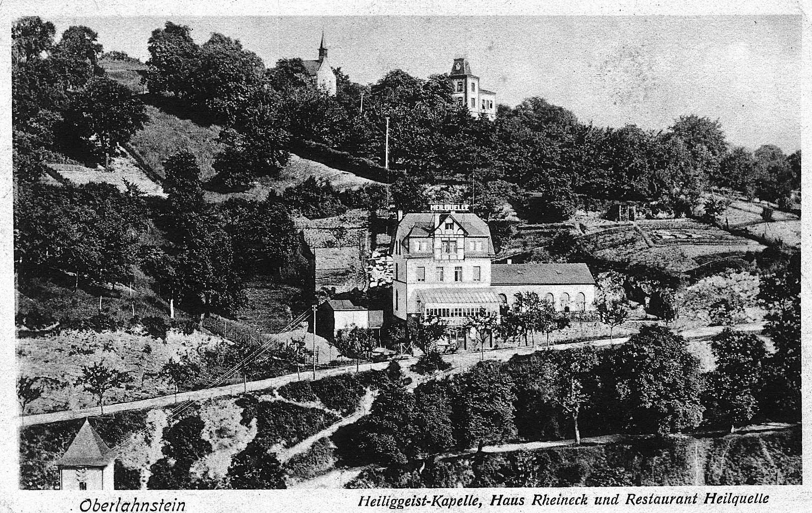 Historische Schwarz-Weiß-Aufnahme von Oberlahnstein. Im Mittelpunkt liegt das Restaurant „Heilquelle“ mit mehreren Gebäudeteilen, umgeben von Gärten, Bäumen und bewachsenen Hängen. Oberhalb sind zwischen den Bäumen die Heiliggeist-Kapelle und das Haus Rheineck zu erkennen. Am unteren Bildrand steht die Beschriftung „Oberlahnstein – Heiliggeist-Kapelle, Haus Rheineck und Restaurant Heilquelle“.