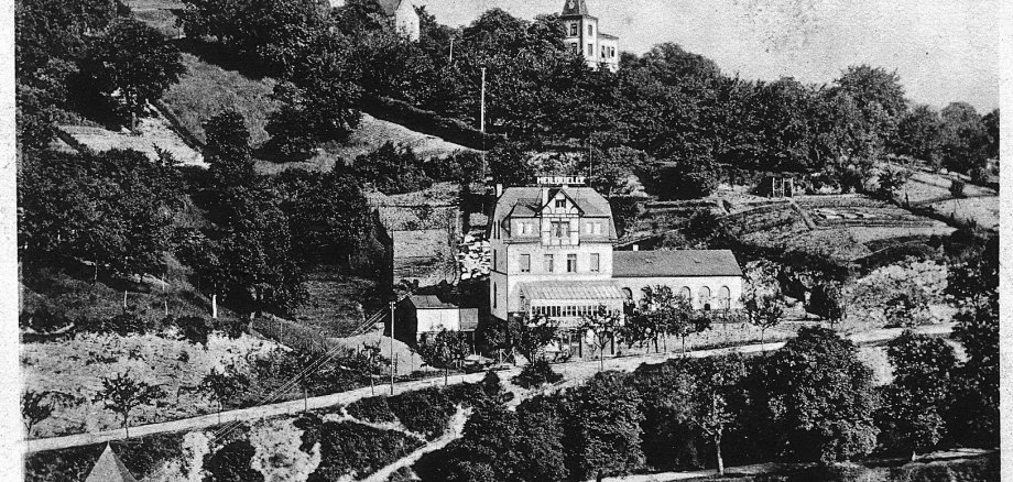 Ansichtskarte um 1910: Oben mittig, gegenüber der Heilig-Geist-Kapelle, steht Haus Rheineck alias Villa Landmann. (Sammlung Stadtarchiv Lahnstein) Historische Schwarz-Weiß-Aufnahme von Oberlahnstein. Im Mittelpunkt liegt das Restaurant „Heilquelle“ mit mehreren Gebäudeteilen, umgeben von Gärten, Bäumen und bewachsenen Hängen. Oberhalb sind zwischen den Bäumen die Heiliggeist-Kapelle und das Haus Rheineck zu erkennen. Am unteren Bildrand steht die Beschriftung „Oberlahnstein – Heiliggeist-Kapelle, Haus Rheineck und Restaurant Heilquelle“.