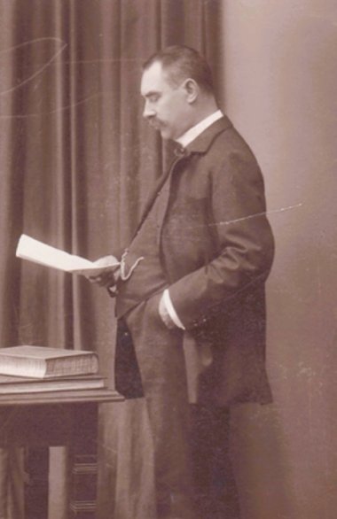 Gottfried Landmann (Sammlung Flohr-Roese) Historische Porträtaufnahme eines Mannes im dunklen Anzug mit Schnurrbart. Er steht seitlich vor einem Vorhang und hält ein Blatt Papier in der linken Hand, auf das er hinabblickt. Die rechte Hand steckt in seiner Hosentasche. Neben ihm steht ein kleiner Tisch mit mehreren Büchern.