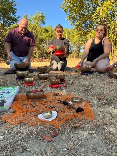 Drei Erwachsene sitzen im Freien auf dem trockenen Waldboden und spielen Klangschalen. Vor ihnen sind auf orangefarbenen und roten Tüchern zahlreiche Klangschalen unterschiedlicher Größe sowie weitere Klanginstrumente angeordnet. Die drei Personen halten jeweils einen Schlägel in der Hand. Im Hintergrund sind Bäume und Sträucher zu sehen; die Aufnahme entstand bei sonnigem Wetter.