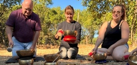 Drei Erwachsene sitzen im Freien auf dem trockenen Waldboden und spielen Klangschalen. Vor ihnen sind auf orangefarbenen und roten Tüchern zahlreiche Klangschalen unterschiedlicher Größe sowie weitere Klanginstrumente angeordnet. Die drei Personen halten jeweils einen Schlägel in der Hand. Im Hintergrund sind Bäume und Sträucher zu sehen; die Aufnahme entstand bei sonnigem Wetter.