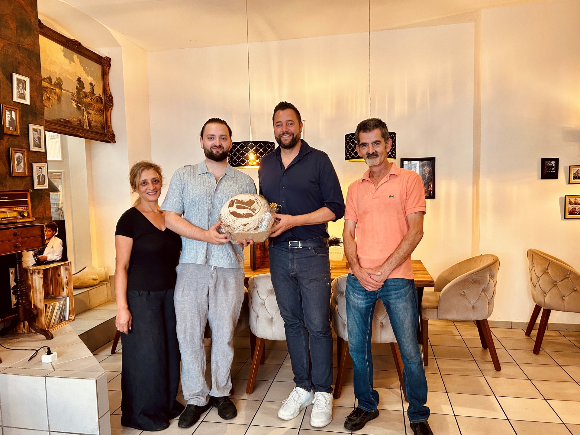 Vier Personen stehen nebeneinander im hell eingerichteten Gastraum des Restaurants Miras in Lahnstein. In der Mitte stehen der Betreiber des Restaurants und Oberbürgermeister Lennart Siefert. Gemeinsam halten sie ein rundes, verpacktes Präsent in den Händen. Links daneben steht eine Frau in schwarzer Kleidung, rechts ein Mann in einem lachsfarbenen Poloshirt. Im Hintergrund sind gedeckte Tische mit gepolsterten Stühlen, schwarze Pendelleuchten sowie zahlreiche gerahmte Bilder an den Wänden zu sehen.