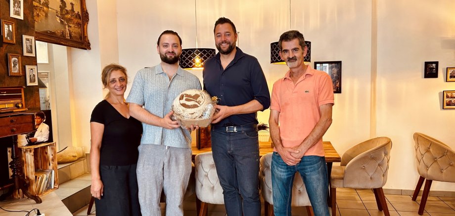 Oberbürgermeister Lennart Siefert (2. v. r.) besuchte das Restaurant Miras in Lahnstein und tauschte sich mit dem Betreiber Sinan Yilmaz (2.v.l.) über das gastronomische Konzept und die weiteren Pläne aus. Vier Personen stehen nebeneinander im hell eingerichteten Gastraum des Restaurants Miras in Lahnstein. In der Mitte stehen der Betreiber des Restaurants und Oberbürgermeister Lennart Siefert. Gemeinsam halten sie ein rundes, verpacktes Präsent in den Händen. Links daneben steht eine Frau in schwarzer Kleidung, rechts ein Mann in einem lachsfarbenen Poloshirt. Im Hintergrund sind gedeckte Tische mit gepolsterten Stühlen, schwarze Pendelleuchten sowie zahlreiche gerahmte Bilder an den Wänden zu sehen.