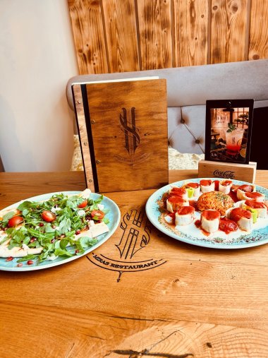 Moderne Fusion Kitchen trifft auf persönliche Wurzeln: Im Miras verbinden sich unterschiedliche kulinarische Einflüsse zu einem besonderen gastronomischen Konzept. Blick auf einen Holztisch im Restaurant Miras mit zwei angerichteten Speisen auf hellblauen Tellern. Links liegt ein belegtes Fladenbrot mit Rucola, Tomaten, Käse und Granatapfelkernen. Rechts sind kleine gefüllte Teigröllchen mit roter Sauce kreisförmig um eine Portion gewürzten Reis angerichtet. Zwischen den Tellern steht die Speisekarte des Restaurants mit einem Holzcover und dem eingravierten Miras-Logo. Das Logo ist auch direkt in die Tischplatte eingearbeitet. Im Hintergrund befindet sich eine gepolsterte Sitzbank vor einer Holzwand.