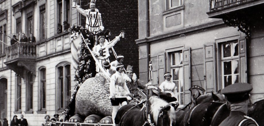 Karnevalsumzug im Jahr 1935 in Lahnstein Historische Schwarz-Weiß-Aufnahme eines Fastnachtsumzugs in Oberlahnstein. Ein von mehreren Pferden gezogener, aufwendig gestalteter Motivwagen fährt durch eine von mehrstöckigen Häusern gesäumte Straße. Auf dem Wagen befinden sich mehrere kostümierte Personen; zwei von ihnen stehen erhöht auf einer großen fantasievollen Figur und grüßen in Richtung der Zuschauer. Weitere Teilnehmer begleiten den Wagen. An den Straßenrändern sowie auf einem Balkon verfolgen Menschen den Umzug. Die Aufnahme vermittelt einen Eindruck von der Lahnsteiner Fastnacht in früheren Jahrzehnten.