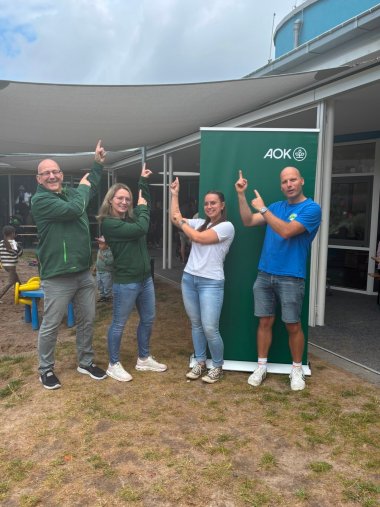 Vier Erwachsene stehen auf dem Außengelände einer Kindertagesstätte nebeneinander vor einem grünen Aufsteller der AOK. Sie lächeln in die Kamera und zeigen mit ausgestreckten Zeigefingern nach oben auf ein großes Sonnensegel über dem Außengelände. Im Hintergrund sind das Kita-Gebäude sowie spielende Kinder zu sehen.