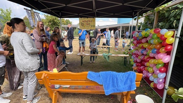 Kinder und Erwachsene beim Sommerfest auf dem Außengelände einer Kindertagesstätte. Unter einem schwarzen Pavillon befindet sich eine Spielstation mit einer großen Wand aus vielen bunten Luftballons. Mehrere Kinder und Erwachsene warten davor oder beobachten das Geschehen. Im Vordergrund stehen eine orangefarbene Holzbank und weitere Utensilien der Spielstation, im Hintergrund sind zahlreiche Besucher des Festes zu sehen.