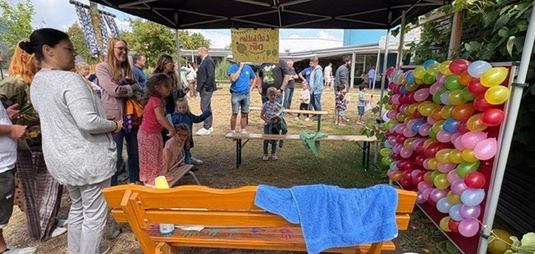 Kinder und Erwachsene beim Sommerfest auf dem Außengelände einer Kindertagesstätte. Unter einem schwarzen Pavillon befindet sich eine Spielstation mit einer großen Wand aus vielen bunten Luftballons. Mehrere Kinder und Erwachsene warten davor oder beobachten das Geschehen. Im Vordergrund stehen eine orangefarbene Holzbank und weitere Utensilien der Spielstation, im Hintergrund sind zahlreiche Besucher des Festes zu sehen.