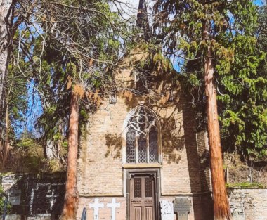 Historische neugotische Gruftkapelle auf dem Friedhof in der Allerheiligenbergstraße in Lahnstein. Die Backsteinfassade wird von einem großen spitzbogigen Fenster mit aufwendigem Maßwerk über dem hölzernen Eingang geprägt. Zwei hohe Bäume rahmen die Kapelle ein, deren Fassade teilweise von Ästen verdeckt wird. Vor dem Gebäude sind mehrere Grabkreuze und Gedenksteine zu sehen.