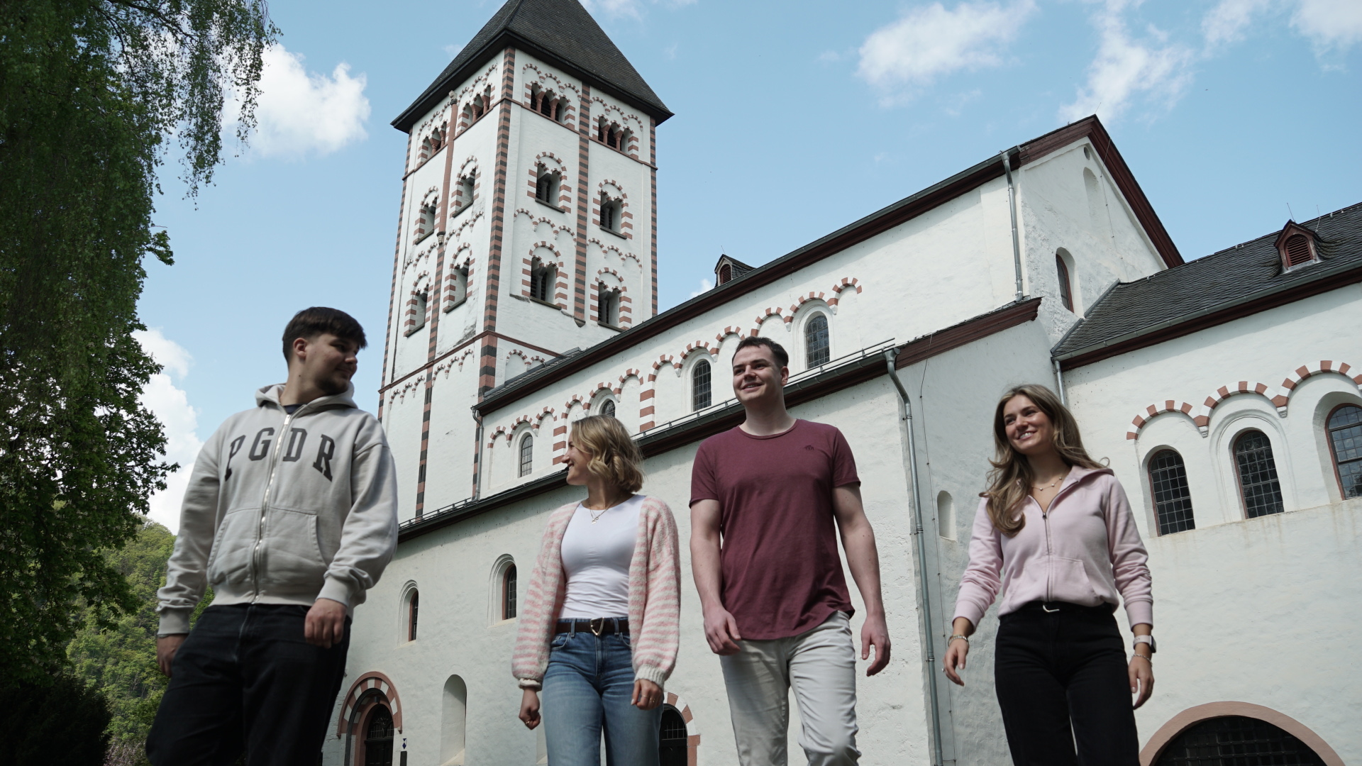 Vier junge Erwachsene gehen vor der historischen Johanniskirche in Lahnstein entlang. Hinter ihnen erhebt sich die weiße Kirche mit ihrem markanten Turm, dunklem Dach und den rot-weiß eingefassten Rundbogenfenstern. Die Aufnahme zeigt die Gruppe bei sonnigem Wetter aus einer niedrigen Perspektive und setzt die romanische Architektur der Kirche deutlich in Szene.