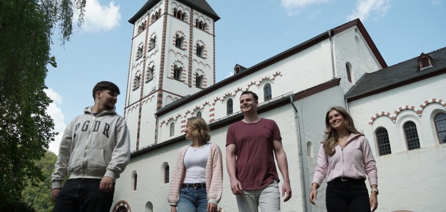 Vier junge Erwachsene gehen vor der historischen Johanniskirche in Lahnstein entlang. Hinter ihnen erhebt sich die weiße Kirche mit ihrem markanten Turm, dunklem Dach und den rot-weiß eingefassten Rundbogenfenstern. Die Aufnahme zeigt die Gruppe bei sonnigem Wetter aus einer niedrigen Perspektive und setzt die romanische Architektur der Kirche deutlich in Szene.