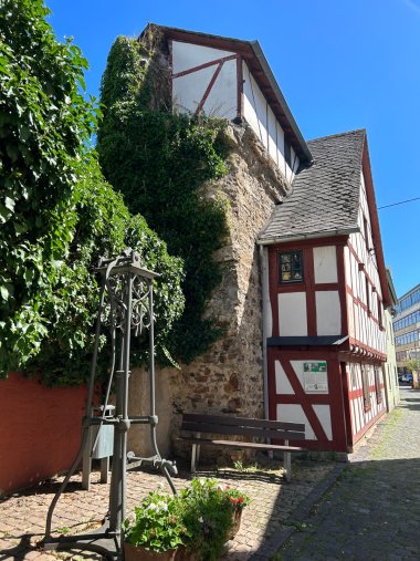 Historisches Stadtmauerhäuschen in Lahnstein bei sonnigem Wetter. Das schmale Gebäude verbindet eine alte, teilweise mit Efeu bewachsene Bruchsteinmauer mit einem weiß und rot gestalteten Fachwerkhaus. Vor dem Gebäude stehen eine Holzbank, ein historisches Metallgestell und ein bepflanzter Kübel. Rechts führt eine gepflasterte Gasse am Häuschen entlang.