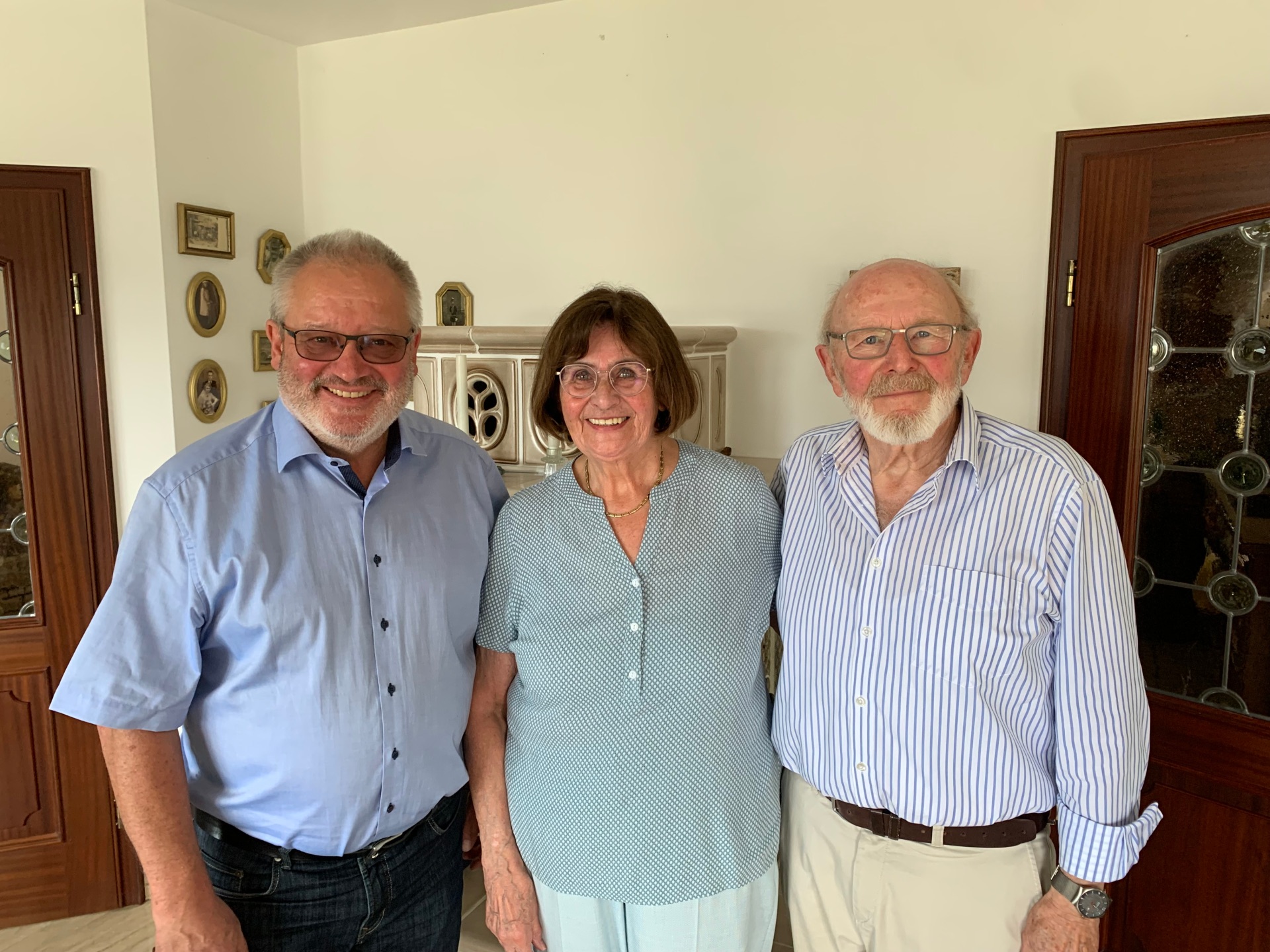Bürgermeister Johannes Lauer steht gemeinsam mit dem Ehepaar Gabi und Theo Dehe in deren Wohnraum. Alle drei blicken lächelnd in die Kamera. Gabi Dehe steht in der Mitte und trägt eine hellgrüne Bluse und eine helle Hose. Rechts neben ihr steht Theo Dehe mit weißem Bart, Brille, blau-weiß gestreiftem Hemd und heller Hose. Links steht Bürgermeister Johannes Lauer mit Brille, hellblauem Hemd und dunkler Hose. Im Hintergrund sind ein heller Kachelofen, mehrere gerahmte Familienbilder sowie zwei Holztüren mit verglasten Einsätzen zu sehen.