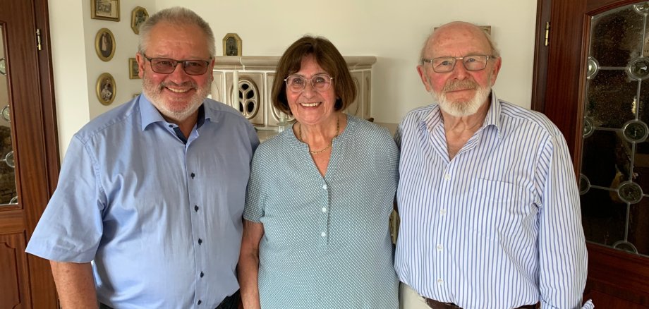 Ehejubiläum Bürgermeister Johannes Lauer steht gemeinsam mit dem Ehepaar Gabi und Theo Dehe in deren Wohnraum. Alle drei blicken lächelnd in die Kamera. Gabi Dehe steht in der Mitte und trägt eine hellgrüne Bluse und eine helle Hose. Rechts neben ihr steht Theo Dehe mit weißem Bart, Brille, blau-weiß gestreiftem Hemd und heller Hose. Links steht Bürgermeister Johannes Lauer mit Brille, hellblauem Hemd und dunkler Hose. Im Hintergrund sind ein heller Kachelofen, mehrere gerahmte Familienbilder sowie zwei Holztüren mit verglasten Einsätzen zu sehen.