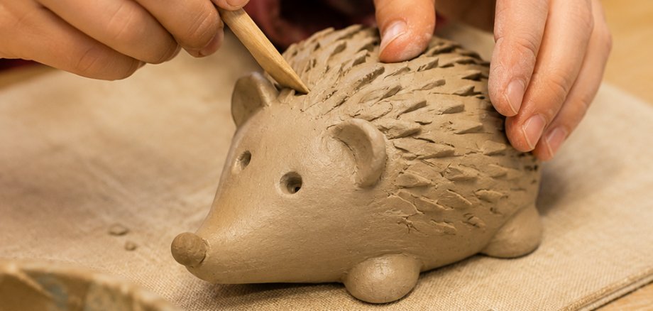 Kinderhände gestalten aus Ton eine Igelfigur. Mit einem Modellierwerkzeug werden die Stacheln in den noch weichen Ton eingearbeitet. Auf dem Tisch liegen weitere Werkzeuge und Tonreste.