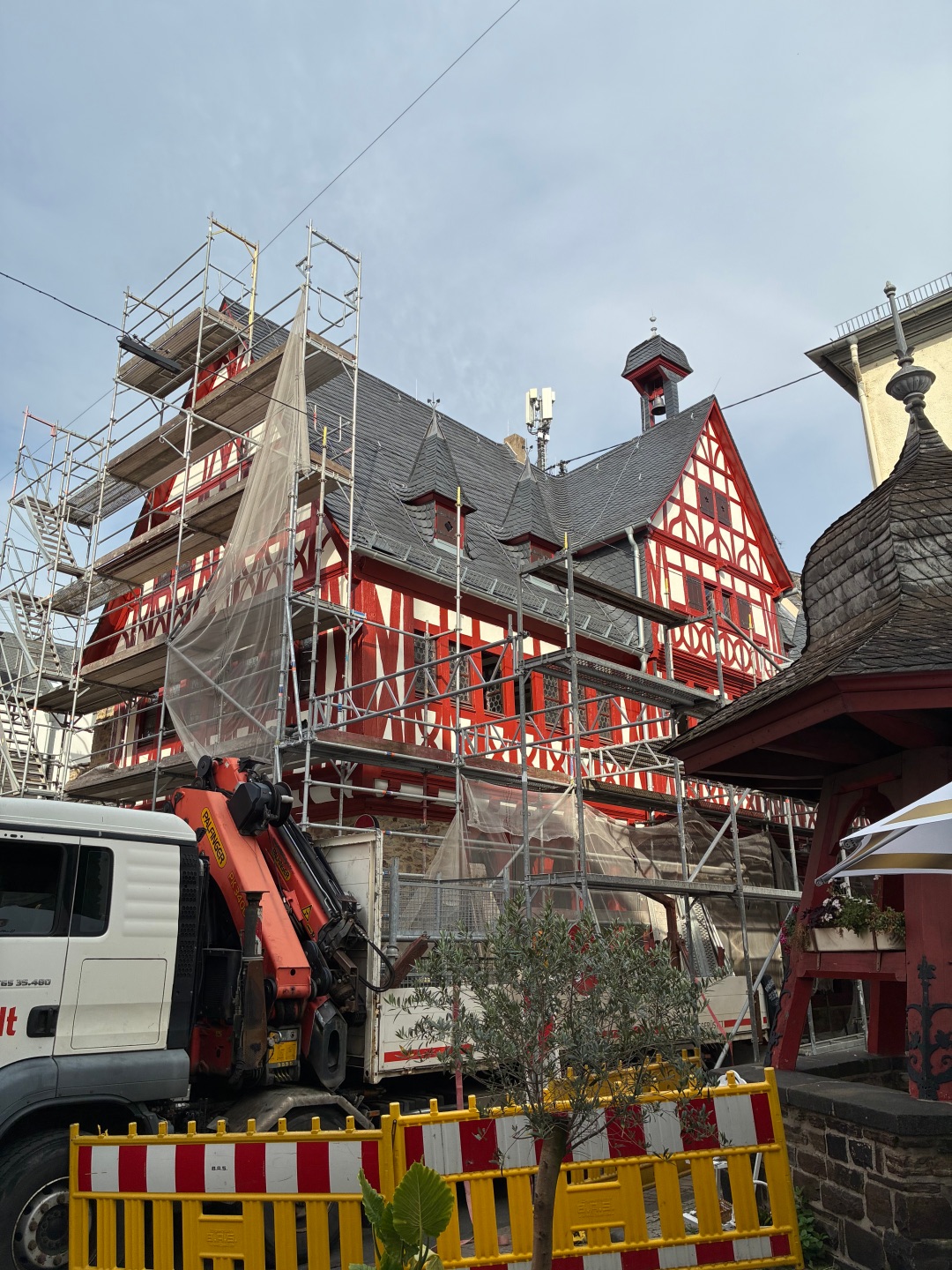 Außenansicht des sanierten Alten Rathauses in Lahnstein mit hälftig abgebautem Gerüst. Die historische Fassade ist geprägt von rot gestrichenem Fachwerk mit hellen Gefachen und einem steinernen Sockelgeschoss. Im Erdgeschoss verlaufen offene, spitzbogige Arkaden entlang des Gebäudes. Mehrere kleine Türmchen und ein Dachreiter mit Glocke ragen aus dem dunkel gedeckten Dach. Das zuvor für die Sanierungsarbeiten aufgestellte Gerüst ist weitgehend abgebaut, sodass die restaurierte Fassade wieder frei sichtbar ist.