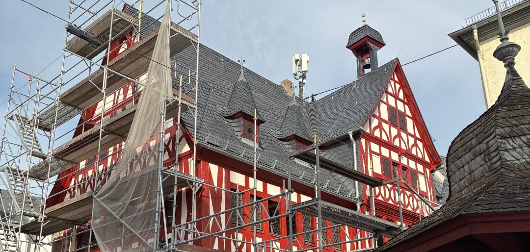 Außenansicht des sanierten Alten Rathauses in Lahnstein mit hälftig abgebautem Gerüst. Die historische Fassade ist geprägt von rot gestrichenem Fachwerk mit hellen Gefachen und einem steinernen Sockelgeschoss. Im Erdgeschoss verlaufen offene, spitzbogige Arkaden entlang des Gebäudes. Mehrere kleine Türmchen und ein Dachreiter mit Glocke ragen aus dem dunkel gedeckten Dach. Das zuvor für die Sanierungsarbeiten aufgestellte Gerüst ist weitgehend abgebaut, sodass die restaurierte Fassade wieder frei sichtbar ist.