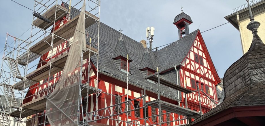Altes Rathaus Lahnstein Außenansicht des sanierten Alten Rathauses in Lahnstein mit hälftig abgebautem Gerüst. Die historische Fassade ist geprägt von rot gestrichenem Fachwerk mit hellen Gefachen und einem steinernen Sockelgeschoss. Im Erdgeschoss verlaufen offene, spitzbogige Arkaden entlang des Gebäudes. Mehrere kleine Türmchen und ein Dachreiter mit Glocke ragen aus dem dunkel gedeckten Dach. Das zuvor für die Sanierungsarbeiten aufgestellte Gerüst ist weitgehend abgebaut, sodass die restaurierte Fassade wieder frei sichtbar ist.