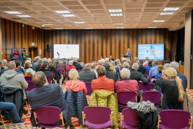 Eine große Gruppe von Personen sitzt in Stuhlreihen mit Blick auf die vorne über Bildschirme gezeigte Präsentation.