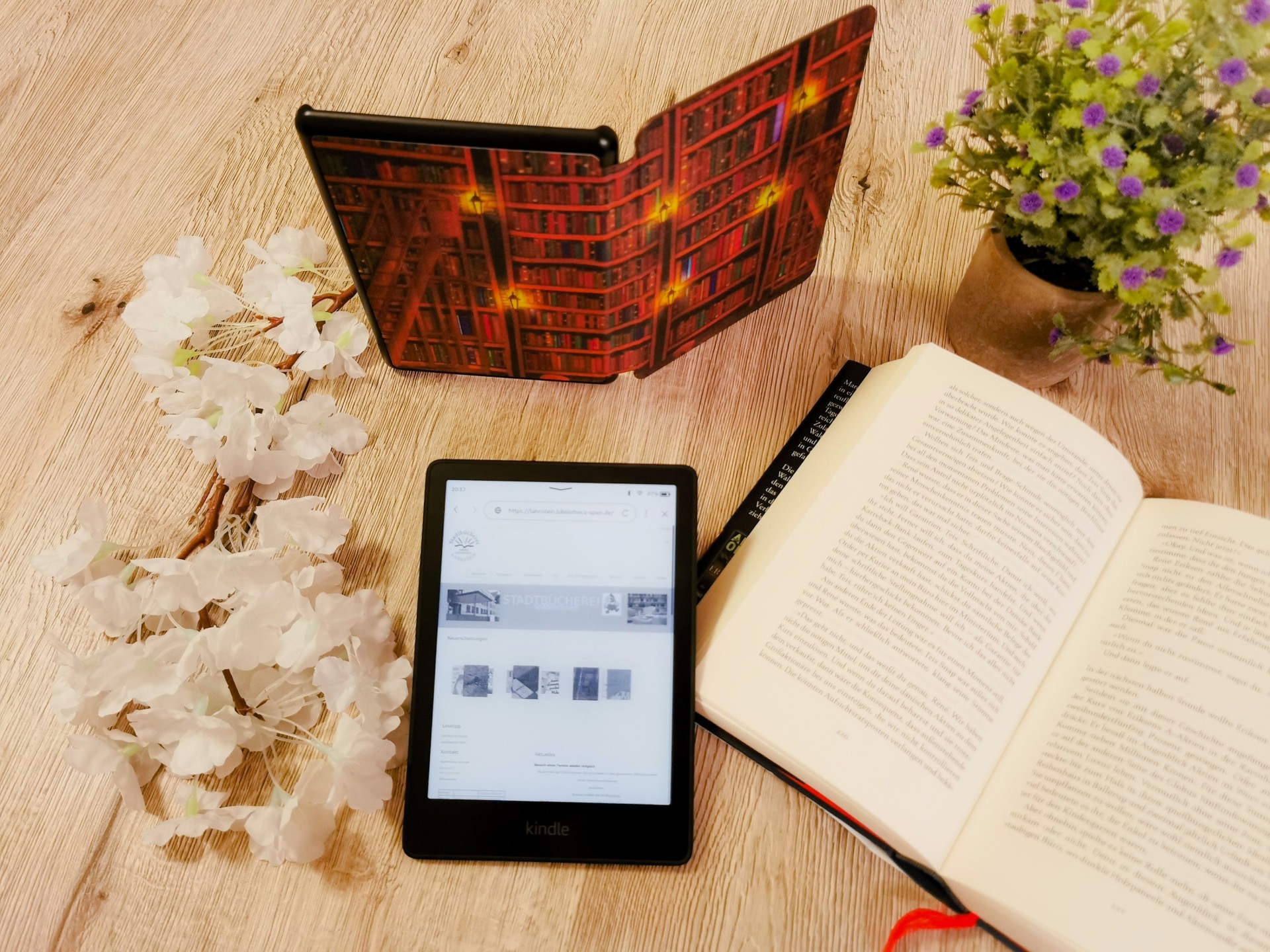Ein eBook-Reader mit Text liegt neben einem aufgeschlagenem Buch, einer Topfpflanze und einem aufgestellten Reader auf einem Tisch.