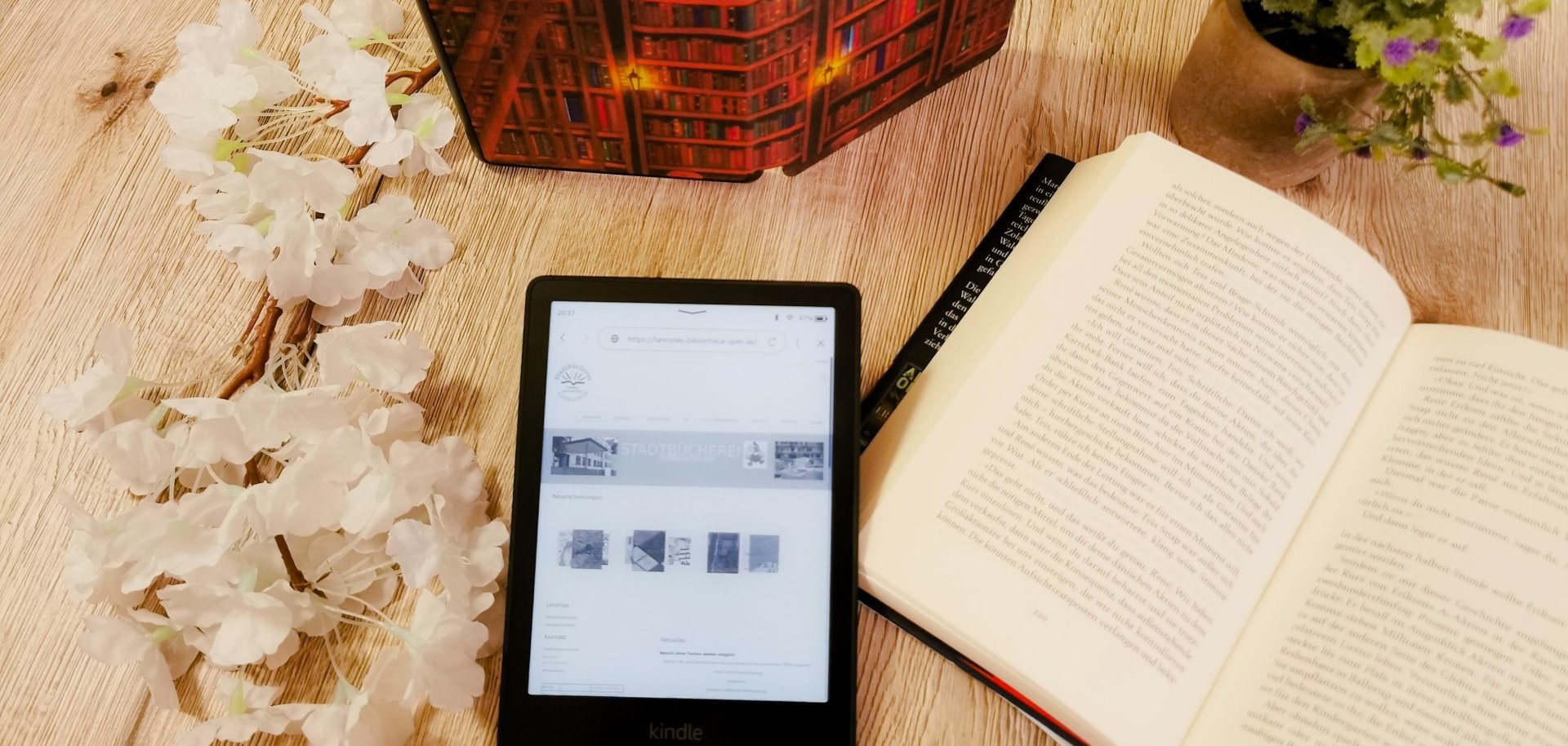 Ein eBook-Reader mit Text liegt neben einem aufgeschlagenem Buch, einer Topfpflanze und einem aufgestellten Reader auf einem Tisch.