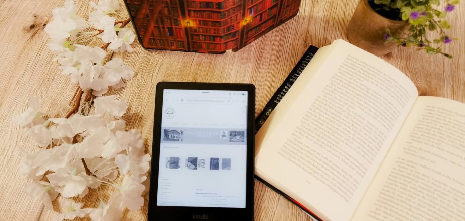 eBook-Sprechstunden in der Stadtbücherei Lahnstein Ein eBook-Reader mit Text liegt neben einem aufgeschlagenem Buch, einer Topfpflanze und einem aufgestellten Reader auf einem Tisch.