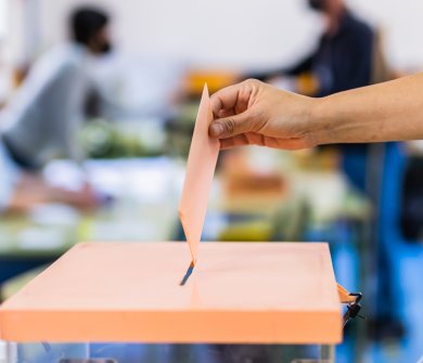 Im Hintergrund sind verschwommen zwei Personen zu erkennen, während im Vordergrund eine durchsichtige Wahlurne steht, in die gerade ein Brief eingeworfen wird. 