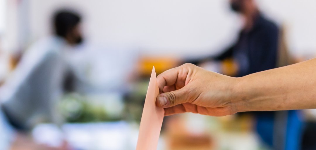 Im Hintergrund sind verschwommen zwei Personen zu erkennen, während im Vordergrund eine durchsichtige Wahlurne steht, in die gerade ein Brief eingeworfen wird. 