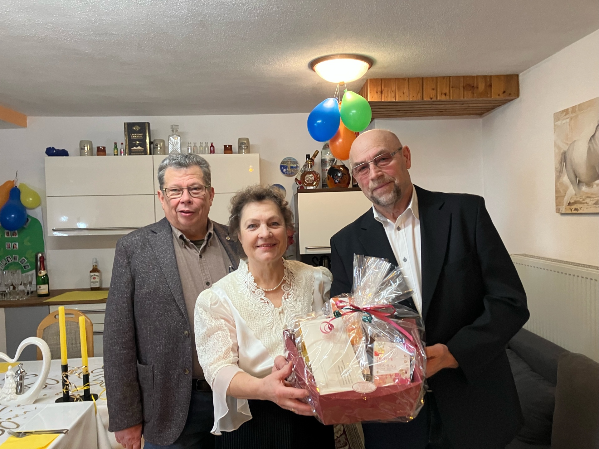 Beigeordneter Manfred Radermacher posiert mit Ehepaar Gallinger für ein Foto. Sie stehen im Haus der Eheleute. Diese halten einen Präsentkorb in die Kamera.