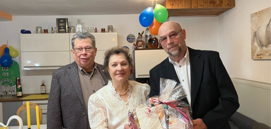 Beigeordneter Manfred Radermacher posiert mit Ehepaar Gallinger für ein Foto. Sie stehen im Haus der Eheleute. Diese halten einen Präsentkorb in die Kamera.