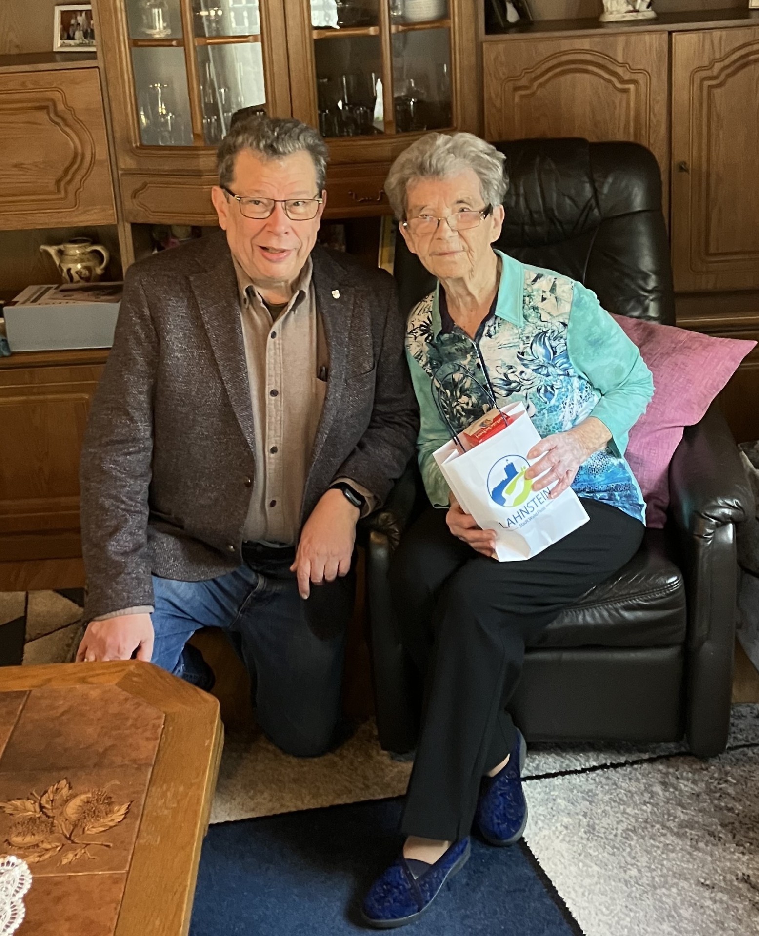 Hilde Rubröder sitzt auf einem Sessel und hält eine mit Lahnstein-Logo bedruckte Tüte in der Hand. Neben ihr hockt Manfred Radermacher.