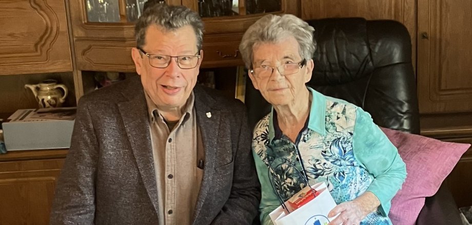 Hilde Rubröder sitzt auf einem Sessel und hält eine mit Lahnstein-Logo bedruckte Tüte in der Hand. Neben ihr hockt Manfred Radermacher.