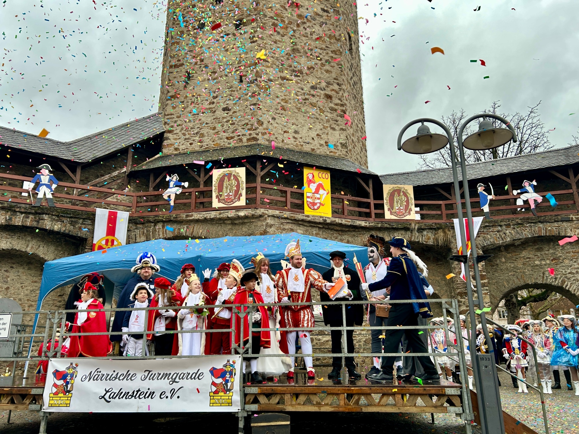 Der Karnevalsprinz steht in Tracht zusammen mit seiner Karnevalsprinzessin und seinem Gefolge sowie mehreren kostümierten Menschen auf einer kleinen Bühne. Konfetti fliegt durch die Luft.