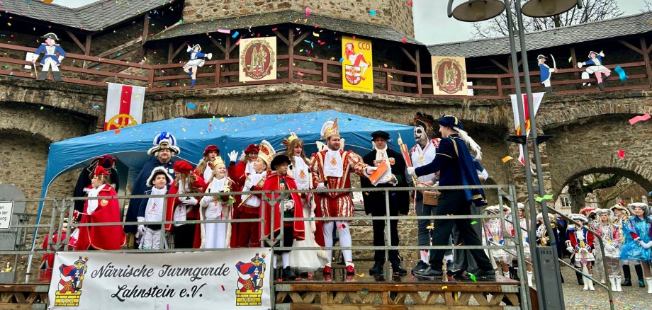 Die Narren erobern Lahnstein. Der Karnevalsprinz steht in Tracht zusammen mit seiner Karnevalsprinzessin und seinem Gefolge sowie mehreren kostümierten Menschen auf einer kleinen Bühne. Konfetti fliegt durch die Luft.