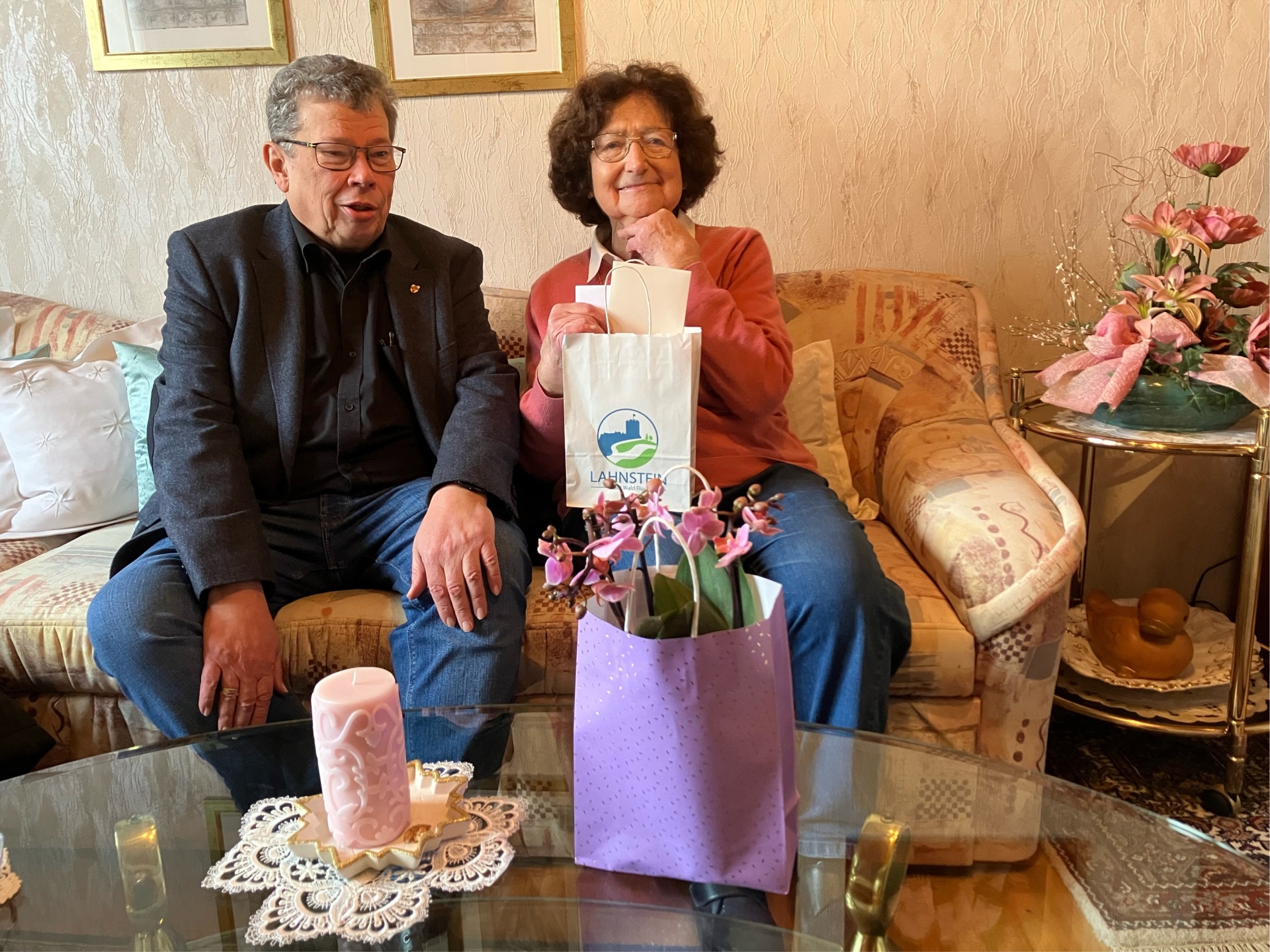 Margot Rick sitzt auf einem Sofa und hält eine mit Lahnstein-Logo bedruckte Tüte in der Hand. Links neben ihr sitzt der Beigeordnete Manfred Radermacher.