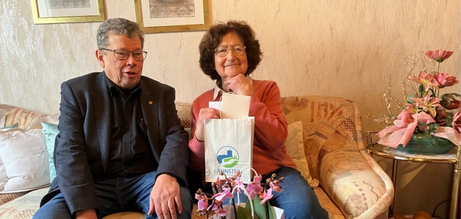 Margot Rick sitzt auf einem Sofa und hält eine mit Lahnstein-Logo bedruckte Tüte in der Hand. Links neben ihr sitzt der Beigeordnete Manfred Radermacher.