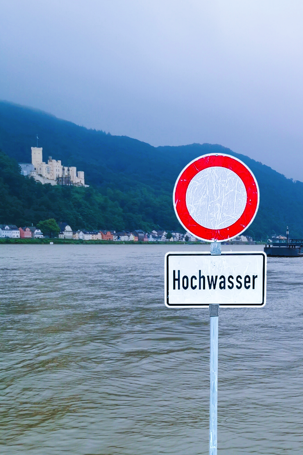 Im Vordergrund steht ein Durchfahrt-verboten-Schild mit dem Zusatzhinweis "Hochwasser". Im Hintergrund ist ein überschwemmter Berech des Rheinufers Oberlahnstein mit Schloss Stolzenfels zu sehen.