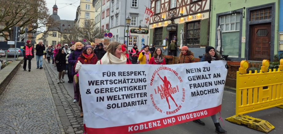Tanz gegen Gewalt: Auch Lahnsteiner Jugendkulturzentrum setzt Zeichen bei „One Billion Rising“ in Koblenz Eine Menge aus überwiegend weiblichen Demonstranten geht gemeinsam auf einer Straße und hält ein Banner hoch, auf dem steht: "Für Respekt, Wertschätzung & Gerechtigkeit in weiltweiter Solidarität" / "Nein zu Gewalt an Frauen und Mädchen"