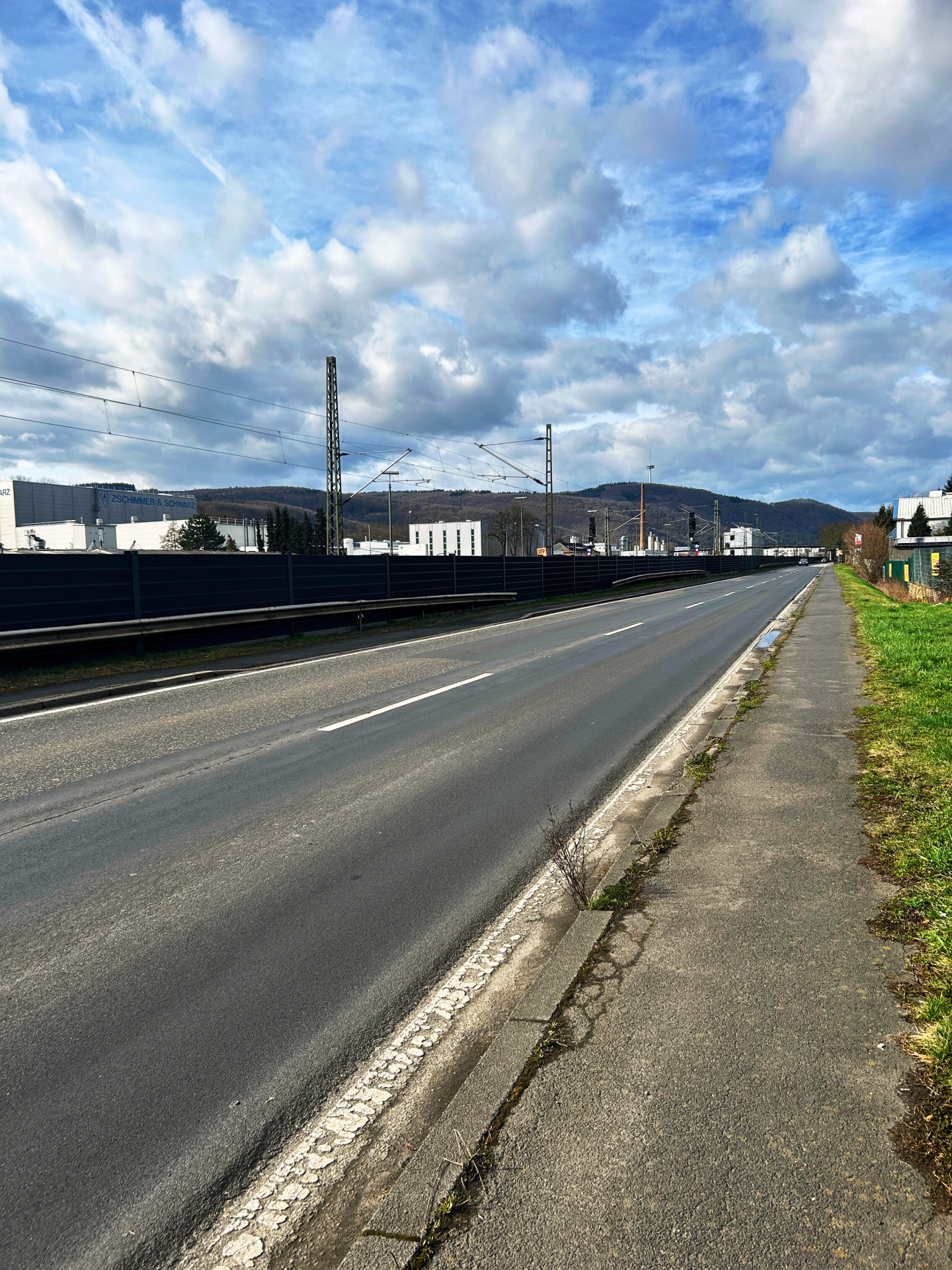 Eine lange Straße, links ist eine Lärmschutzwand und rechts sieht man etwas Gehweg mit begrüntem Randstreifen erkennbar