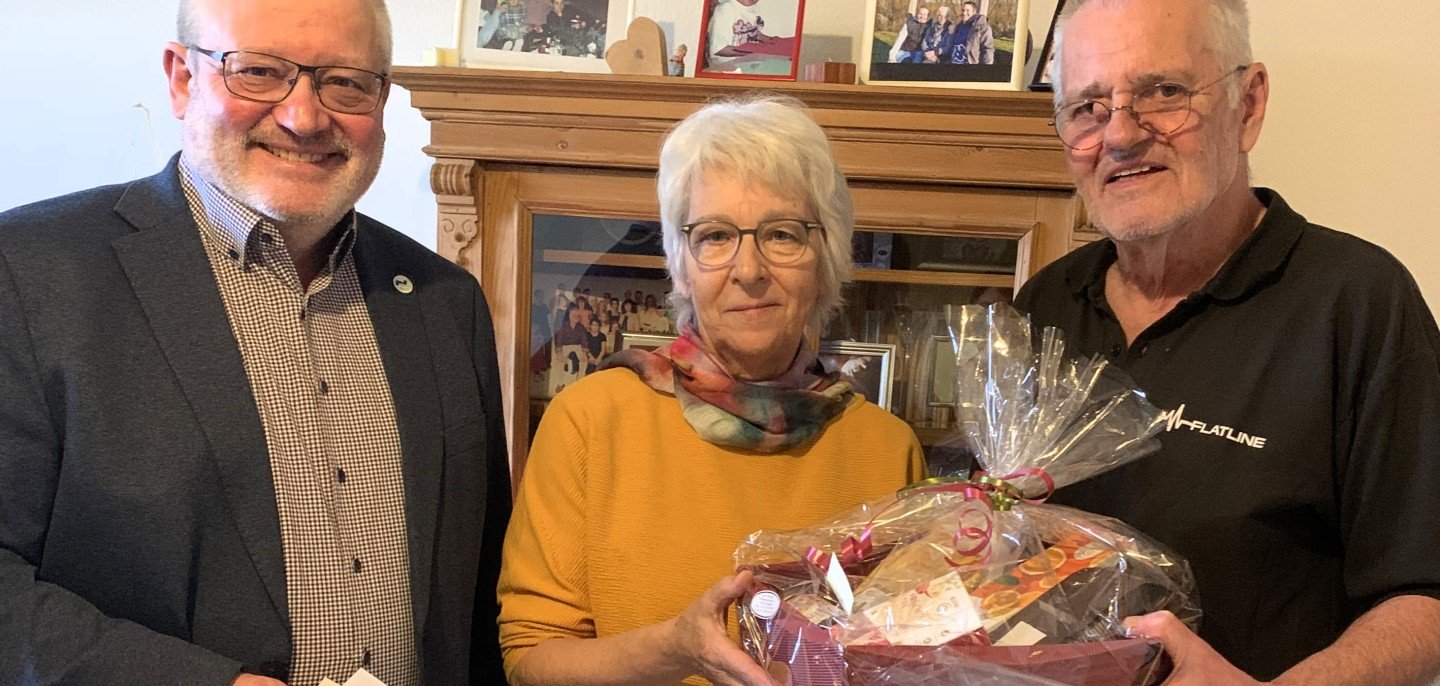 Bürgermeister Johannes Lauer steht neben dem Jubiläumspaar Karin und Wolfgang Michael. Die beiden halten zusammen einen Präsentkorb in den Händen. Im Hintergrund sind Bilderrahmen auf einem Schrank zu erkennen.