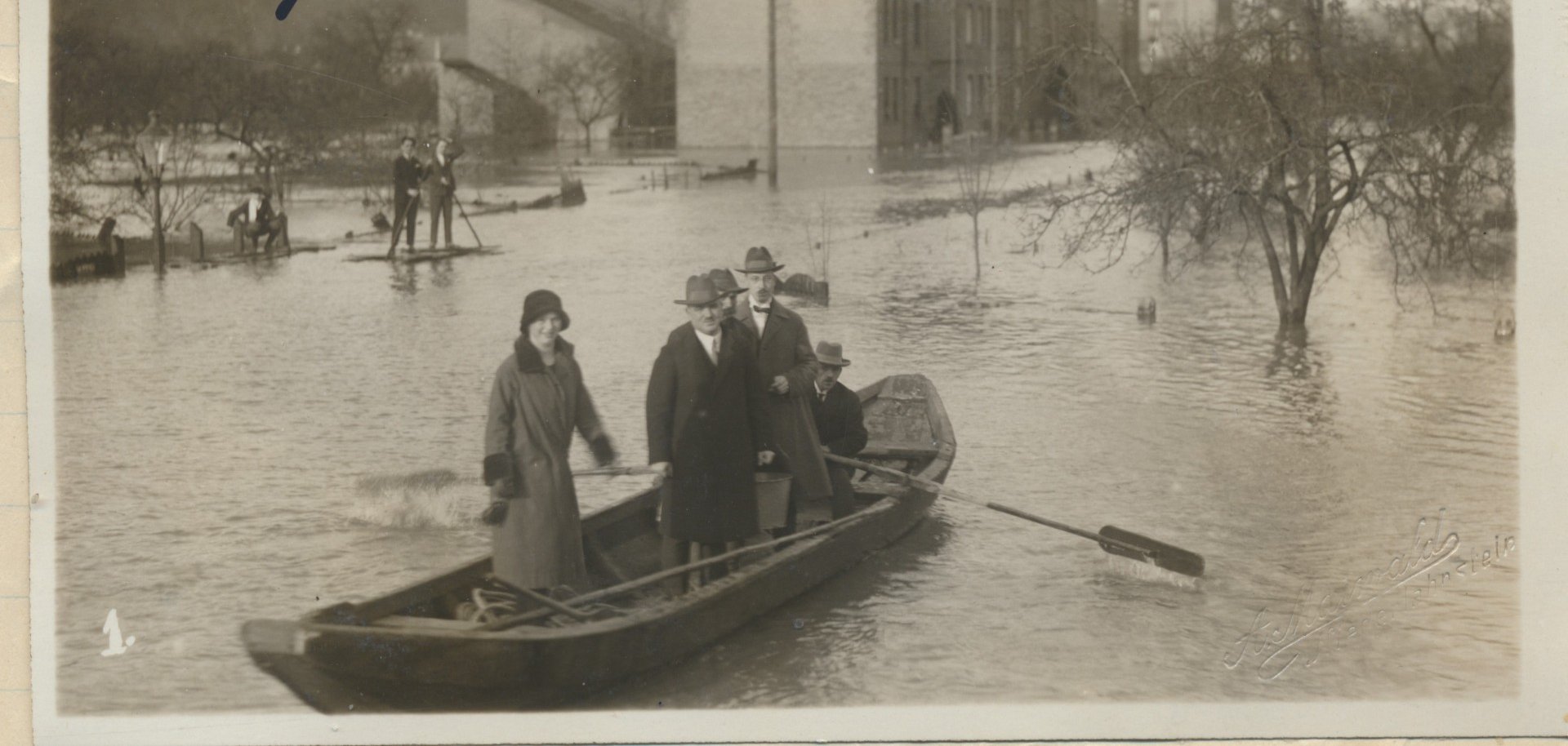 Ein sepiafarbenes Foto aus dem Jahr 1926. Es ist Hochwasser in Lahnstein und Menschen fahren mit einem kleinen Ruderboot durch das überschwemmte Gebiet. Im Hintergrund sind Häuser im Wasser und zwei Männer auf einem Floß erkennbar.