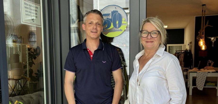 30. Jubiläum von „Ulli b. Lifestyle“ in Lahnstein Ein Mann und eine Frau stehen lächelnd im Eingangsbereich eines Geschäfts. Hinter ihnen ist ein heller Ladenraum mit Kleidung und Dekoration zu sehen. Beide wirken entspannt und freundlich, während sie in die Kamera schauen. Hinter dem Kopf des Mannes ist ein großer, runder Aufkleber zu sehen. Er ist in den Farben Grün und Blau gestaltet und zeigt eine große 30.