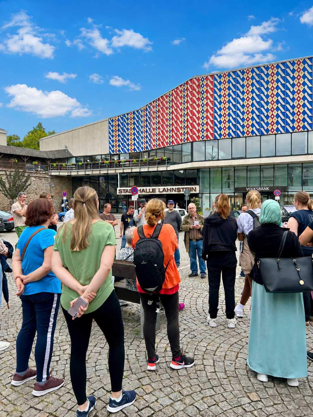 Eine Gruppe von Teilnehmerinnen und Teilnehmern steht auf einem gepflasterten Platz, dem Salhofplatz, vor der Stadthalle Lahnstein und lauscht einem Stadtführer. Im Hintergrund ist die markante Fassade der Stadthalle mit ihrem rot-blauen Muster zu sehen. Die Menschen stehen in lockerer Runde beisammen, während leichte Wolken bei blauem Himmel über dem Gebäude hängen. Das Bild zeigt eine geführte Tour in Lahnstein.