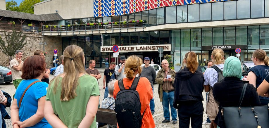 Stadtführung Lahnstein Eine Gruppe von Teilnehmerinnen und Teilnehmern steht auf einem gepflasterten Platz, dem Salhofplatz, vor der Stadthalle Lahnstein und lauscht einem Stadtführer. Im Hintergrund ist die markante Fassade der Stadthalle mit ihrem rot-blauen Muster zu sehen. Die Menschen stehen in lockerer Runde beisammen, während leichte Wolken bei blauem Himmel über dem Gebäude hängen. Das Bild zeigt eine geführte Tour in Lahnstein.
