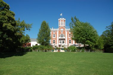 Ein historisches Schloss mit roter Backsteinfassade, weißen Zierelementen und einem zentralen Turm steht inmitten eines weitläufigen Parks. Vor dem Gebäude erstreckt sich eine große Rasenfläche, die von Bäumen und Sträuchern eingerahmt wird. Auf der Terrasse vor dem Schloss stehen Tische, Stühle und Sonnenschirme. Über dem Turm weht eine Flagge vor einem wolkenlosen blauen Himmel.