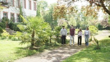 Vier junge Erwachsene gehen gemeinsam auf einem geschwungenen Weg durch einen parkähnlichen Garten mit Palmen und großen Laubbäumen. Links steht ein historisches Gebäude mit heller Fassade und hohen Fenstern. Die Gruppe unterhält sich beim Spaziergang, während im Hintergrund weitläufige Grünflächen und blühende Bäume zu sehen sind.