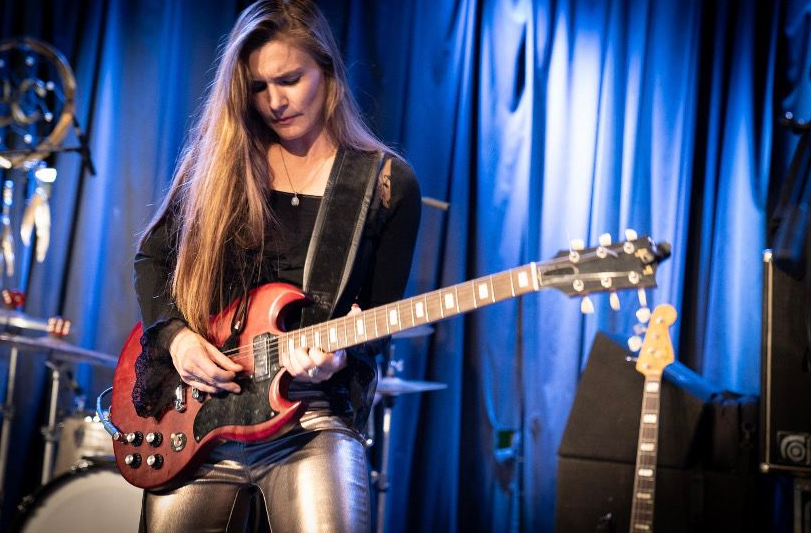 Eine Gitarristin spielt konzentriert ein Solo auf einer rotbraunen E-Gitarre. Sie trägt ein schwarzes Oberteil und silberglänzende Hosen. Die Bühne ist in blaues Licht getaucht, im Hintergrund sind ein Schlagzeug sowie weitere Instrumente zu erkennen. Die Aufnahme fängt einen intensiven Moment eines Live-Konzerts ein.