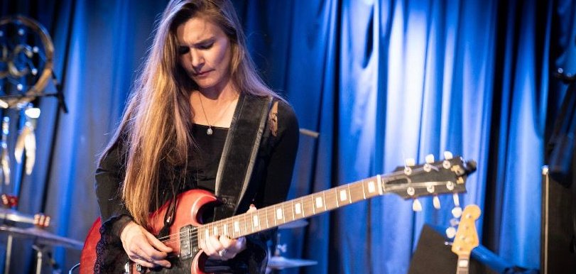 Eine Gitarristin spielt konzentriert ein Solo auf einer rotbraunen E-Gitarre. Sie trägt ein schwarzes Oberteil und silberglänzende Hosen. Die Bühne ist in blaues Licht getaucht, im Hintergrund sind ein Schlagzeug sowie weitere Instrumente zu erkennen. Die Aufnahme fängt einen intensiven Moment eines Live-Konzerts ein.