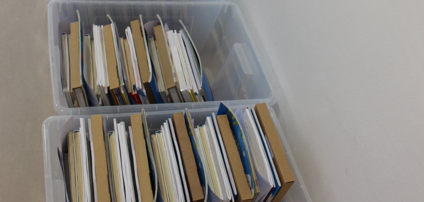 Mehrere transparente Kunststoffboxen mit nach Klassen sortierten Schulbuchpaketen stehen in einer langen Reihe entlang einer weißen Wand in der Stadtbücherei Lahnstein.